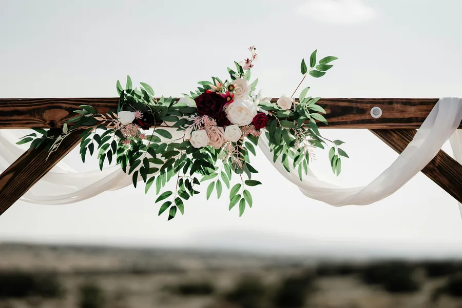 A wooden ceremony arch dressed with roses, greenery and draped fabric above desert scrub
