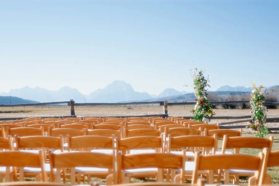 Rows of wooden ceremony chairs facing two floral columns, a split-rail fence and dry open range with mountains beyond