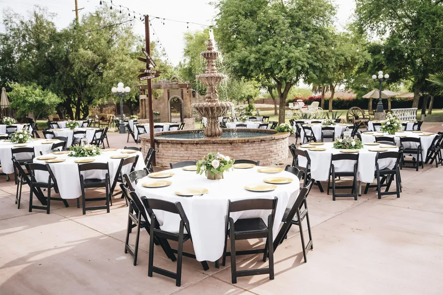 Round reception tables with white linens and gold chargers arranged around a stone fountain on a patio